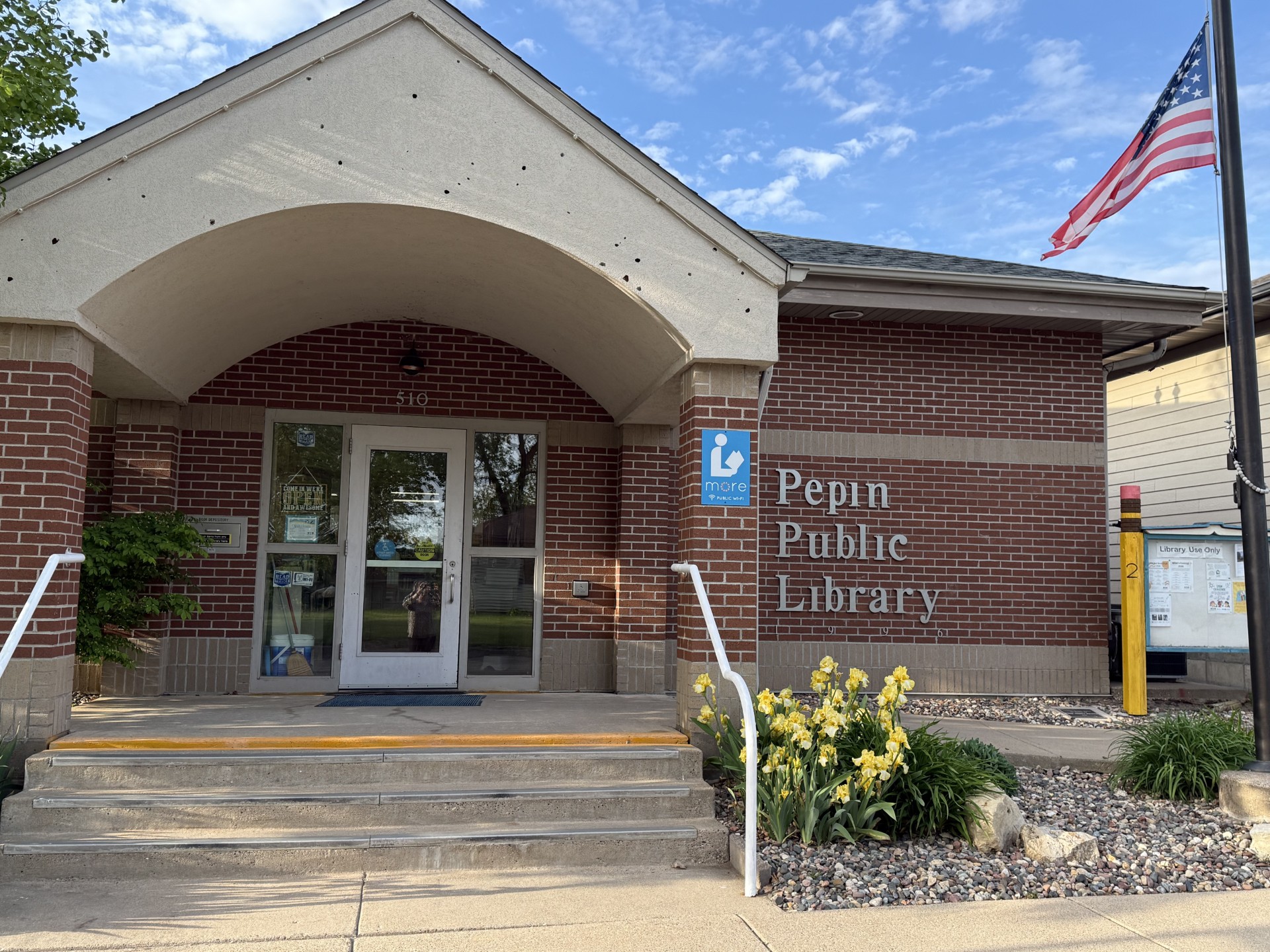 exterior of the pepin public library a red brick building with yellow irises in front and a flag on a flag pole
